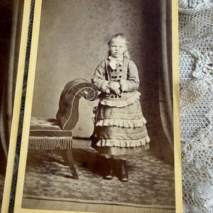 May include: Sepia-toned portrait of a young girl in a dress with layered ruffles, standing next to a decorative bench. The girl wears a bow and buttoned jacket. The photograph is mounted on a vintage card.