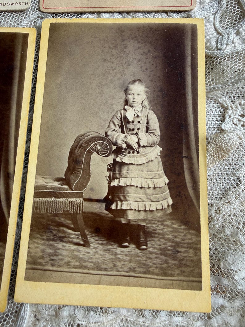 May include: Sepia-toned portrait of a young girl in a dress with layered ruffles, standing next to a decorative bench. The girl wears a bow and buttoned jacket. The photograph is mounted on a vintage card.