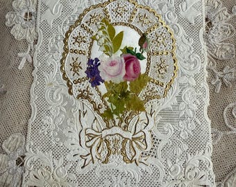 Puede incluir: Una tarjeta de encaje blanco con un óvalo dorado que presenta un ramo de flores rosas y moradas. La tarjeta tiene las palabras "A Whisper of Love" escritas en ella.