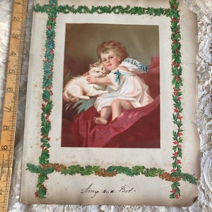 May include: An antique Christmas card featuring a young girl in a white dress holding a white kitten. The card is decorated with a green and red holly border and the date "Aug. 4th 1897" is written in cursive.