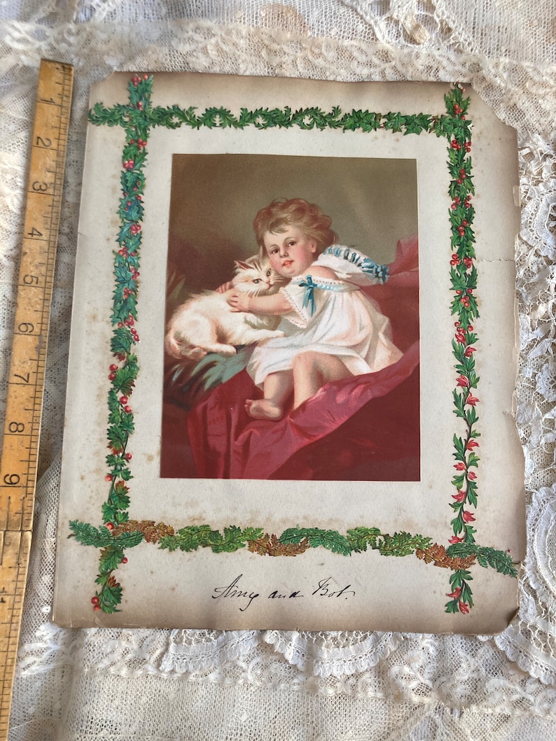 May include: An antique Christmas card featuring a young girl in a white dress holding a white kitten. The card is decorated with a green and red holly border and the date "Aug. 4th 1897" is written in cursive.