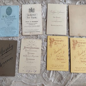 May include: A collection of vintage photographic studio cards. The cards vary in color, including blue, tan, and yellow. Several cards feature text, including studio names, addresses, and the names of photographers. The cards are arranged on a textured surface.