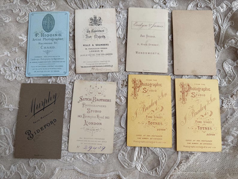 May include: A collection of vintage photographic studio cards. The cards vary in color, including blue, tan, and yellow. Several cards feature text, including studio names, addresses, and the names of photographers. The cards are arranged on a textured surface.