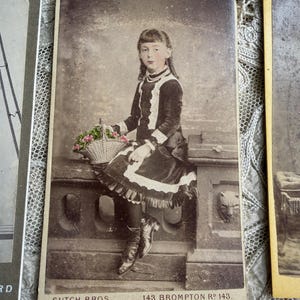 May include: Vintage sepia-toned photograph of a young girl seated on a stone ledge, holding a basket of flowers. She wears a dark dress with white lace trim, a pearl necklace, and boots. The photograph is labeled "SUTCH BROS, 143 BROMPTON Rd 143."