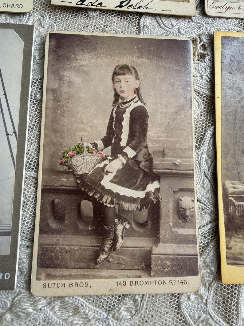May include: Vintage sepia-toned photograph of a young girl seated on a stone ledge, holding a basket of flowers. She wears a dark dress with white lace trim, a pearl necklace, and boots. The photograph is labeled "SUTCH BROS, 143 BROMPTON Rd 143."