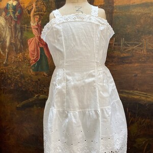 May include: White cotton slip dress with delicate eyelet lace trim and a tiered skirt. The dress has a fitted bodice and a gathered waist.