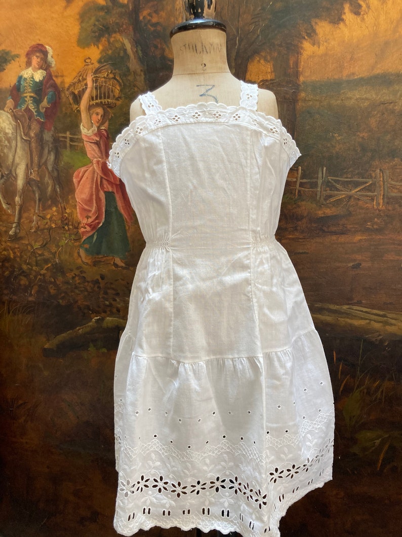 May include: White cotton slip dress with delicate eyelet lace trim and a tiered skirt. The dress has a fitted bodice and a gathered waist.