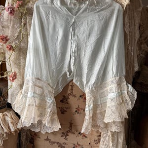 May include: A pale blue blouse featuring a high neckline and long, wide sleeves that end in tiered, cream-colored lace ruffles. The garment is constructed from a lightweight, slightly wrinkled fabric and is displayed on a white hanger.