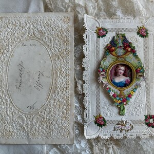 May include: Antique greeting cards. One card features an oval portrait of a woman in a blue dress, surrounded by floral and fruit embellishments. The other card has an embossed floral design with handwritten text.