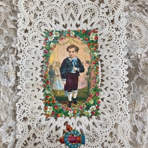 May include: A vintage lace doily with a colorful illustration of a young boy in a blue jacket and red pants standing in a garden setting. The illustration is surrounded by a border of red berries and white flowers. The doily has a delicate, intricate design.