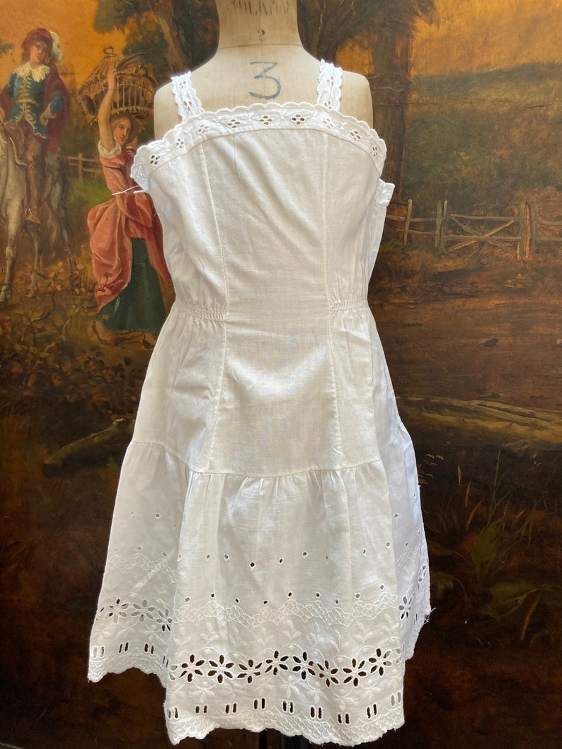May include: White cotton slip dress with eyelet lace trim and a tiered skirt. The dress is on a mannequin and has a vintage look.