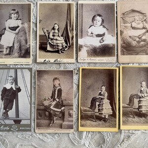 May include: A collection of eight antique cabinet cards featuring portraits of children. The sepia-toned photographs showcase children in various poses, including standing and seated, with detailed clothing and backgrounds. The cards are arranged on a lace surface.