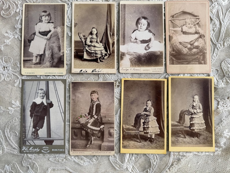 May include: A collection of eight antique cabinet cards featuring portraits of children. The sepia-toned photographs showcase children in various poses, including standing and seated, with detailed clothing and backgrounds. The cards are arranged on a lace surface.