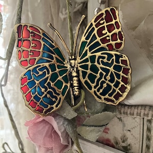 May include: A gold-colored butterfly ornament with red, blue, and green enamel detailing. The butterfly is perched on a stem with a pink flower.
