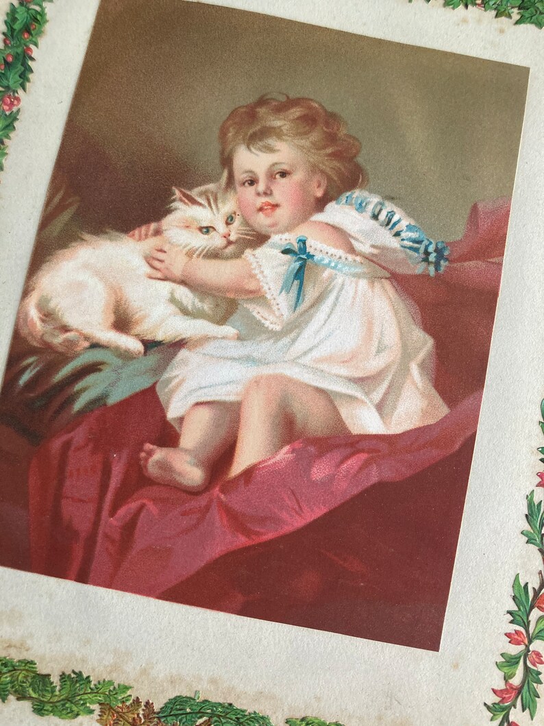 May include: A vintage illustration of a young girl in a white dress holding a white cat. The girl is sitting on a red velvet cushion and the cat is resting on her lap. The illustration is framed with a green floral border.