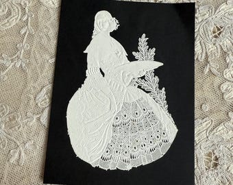May include: A white paper cut-out of a woman in a long dress, holding a tray, set against a black background. The dress features intricate lace-like detailing. The artwork is displayed on a textured, off-white surface.