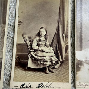 May include: Sepia-toned antique photograph of a young girl seated in a chair. She wears a dress with a ruffled skirt and a bow in her hair. The photograph is labeled "- Ada Deleh -" in cursive.