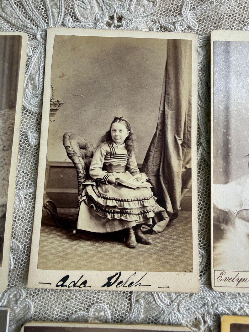 May include: Sepia-toned antique photograph of a young girl seated in a chair. She wears a dress with a ruffled skirt and a bow in her hair. The photograph is labeled "- Ada Deleh -" in cursive.