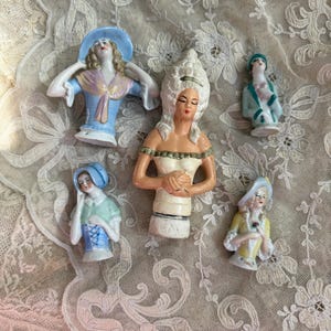 May include: Five vintage porcelain doll heads with different hairstyles and clothing. The dolls are wearing blue, green, yellow, and white clothing. The dolls are all in various poses.