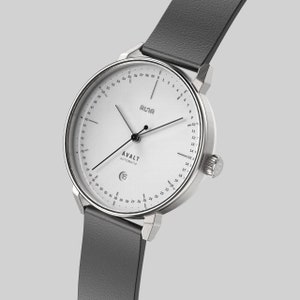 Può includere: Un orologio da polso argentato con quadrante bianco e lancette nere. L'orologio ha una finestrella per la data alle 3 e una scala di tempo di 24 ore attorno al bordo esterno del quadrante. L'orologio è su un cinturino in pelle nera.