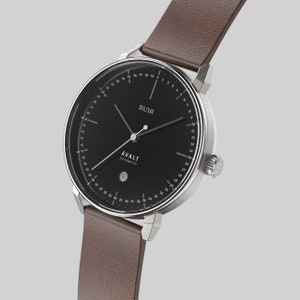 Könnte beinhalten: Eine silberne Uhr mit schwarzem Zifferblatt und braunem Lederarmband. Das Zifferblatt der Uhr hat ein Datumsfenster und die Wörter "RLIA" und "AVALT AUTOMATIC" sind darauf gedruckt.