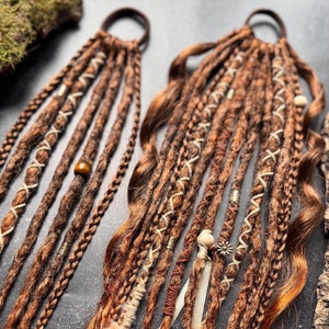 May include: Two sets of brown synthetic dreadlocks with white and brown accents. Each set is tied together with a brown elastic band. The dreadlocks are decorated with beads and a small wooden charm.