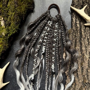 Op de afbeelding: Een set zwarte en witte haar extensions met een bruin elastiek. De extensions zijn gevlochten en hebben witte stiksels en zwarte kralen.