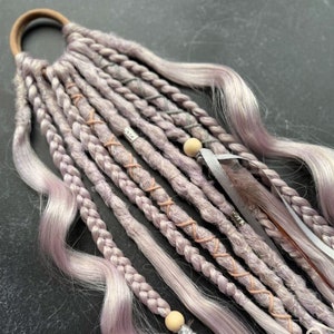 Op de afbeelding: Een set van lavendel en grijze synthetische haarverlengingen met kralen, veren en lint accenten. De extensions zijn gevlochten en hebben een golvende textuur.