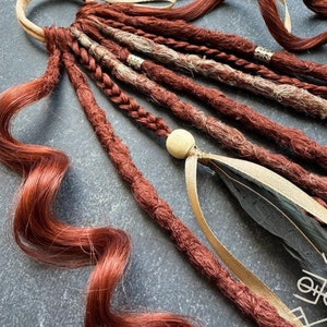 Pode incluir: Um conjunto de dreadlocks sintéticas vermelhas e marrons com detalhes dourados e brancos. As dreadlocks são amarradas com uma faixa de couro marrom e uma fita dourada. Uma pena preta está presa na parte inferior das dreadlocks.