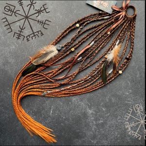 May include: A set of brown hair extensions featuring multiple braided strands attached to a circular ring. The extensions are decorated with brown feathers, silver-toned metal beads, wooden beads, and a brown ribbon tied in a bow at the top.