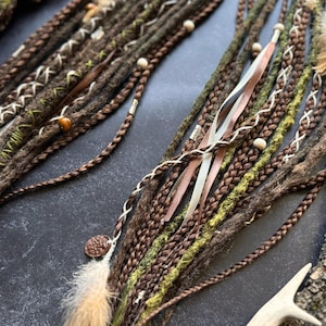 Op de afbeelding: Bruine, groene en witte synthetische dreadlocks met kralen, veren en lint accenten.