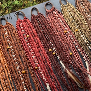 Puede incluir: Una variedad de extensiones de rastas coloridas en tonos marrón, rojo y dorado. Cada rasta está adornada con costuras blancas y cuentas de madera. Algunas extensiones presentan plumas. Las rastas están unidas a gomas para el pelo marrones.