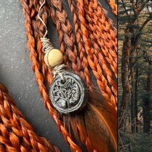 May include: Close-up of a set of braided hair extensions in a reddish-brown color. The extensions are adorned with a small wooden bead and a charm featuring a tree design. The charm is attached to a brown feather.
