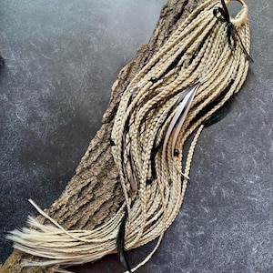 May include: A set of light beige synthetic hair extensions featuring multiple thin braids. The hair is accented with black ribbons, small black beads, and long, thin black and white feathers attached to the strands, creating a textured hair accessory.