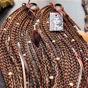 May include: Brown braided hair extensions with white beads, brown leather cords, and a small feather accent. The extensions are tied together with a brown leather cord.