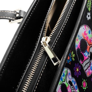 May include: Close-up of a black tote bag with a gold zipper. The bag has a colorful sugar skull print.