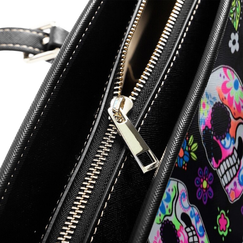 May include: Close-up of a black tote bag with a gold zipper. The bag has a colorful sugar skull print.