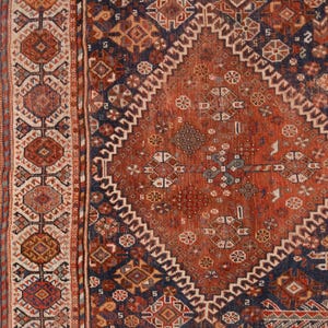 May include: Close-up of a section of an antique rug with a detailed geometric pattern. The rug features a central diamond design in shades of rust and navy blue, surrounded by intricate borders with repeating motifs. The rug's texture and color palette suggest a vintage or traditional style.