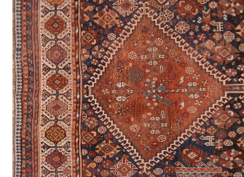 May include: Close-up of a section of an antique rug with a detailed geometric pattern. The rug features a central diamond design in shades of rust and navy blue, surrounded by intricate borders with repeating motifs. The rug's texture and color palette suggest a vintage or traditional style.