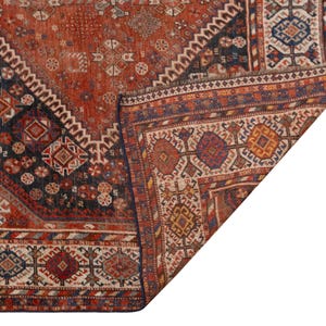 May include: A section of a vintage rug with a detailed geometric pattern. The rug features a rich color palette of rust, navy, and cream, with intricate diamond and floral motifs. The corner is folded over, revealing the rug's construction.