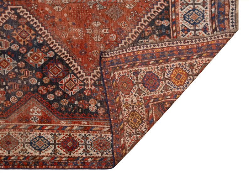 May include: A section of a vintage rug with a detailed geometric pattern. The rug features a rich color palette of rust, navy, and cream, with intricate diamond and floral motifs. The corner is folded over, revealing the rug's construction.