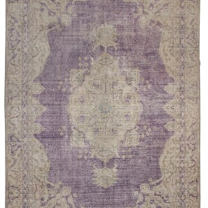 K&ouml;nnte beinhalten: Ein Vintage-Teppich mit einem violetten Hintergrund und einem beigefarbenen Muster. Der Teppich hat ein zentrales Medaillon mit komplizierten Details und einen Rand mit floralen Motiven.