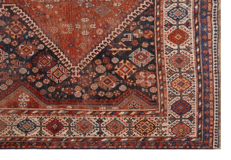 May include: Close-up of a section of an antique rug with a geometric pattern. The rug features a central diamond design in shades of red and navy blue, with intricate borders in cream, red, and blue. The rug's design is detailed and complex.