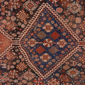 May include: Close-up of a detailed, patterned rug with a geometric design. The rug features a central diamond shape in deep blue, surrounded by red and brown geometric patterns. The edges are bordered with a white zig-zag pattern.