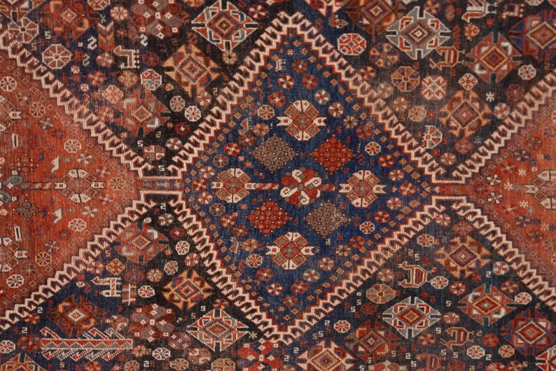 May include: Close-up of a detailed, patterned rug with a geometric design. The rug features a central diamond shape in deep blue, surrounded by red and brown geometric patterns. The edges are bordered with a white zig-zag pattern.