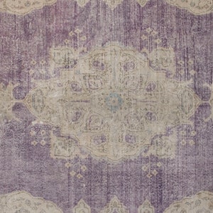 K&ouml;nnte beinhalten: Ein Vintage-Teppich mit einem verblassten, komplizierten Design in Violett- und Beiget&ouml;nen. Der Teppich hat ein zentrales Medaillon mit einem Blumenmuster und einen Rand mit sich wiederholenden Motiven.