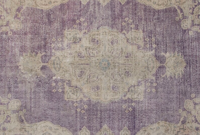 K&ouml;nnte beinhalten: Ein Vintage-Teppich mit einem verblassten, komplizierten Design in Violett- und Beiget&ouml;nen. Der Teppich hat ein zentrales Medaillon mit einem Blumenmuster und einen Rand mit sich wiederholenden Motiven.