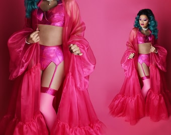 May include: A person wearing a pink satin robe with a ruffled hem and a matching lingerie set. The lingerie set includes a bra, a high-waisted panty, and a garter belt. The person has teal blue hair and is standing on a pink background.