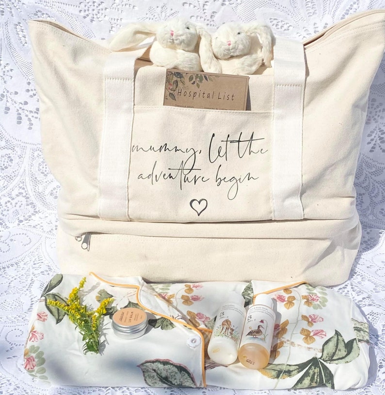 Op de afbeelding: Een witte canvas draagtas met de tekst "mummy, let the adventure begin" en een hart. De tas is gevuld met een ziekenhuislijst, twee witte konijnenknuffels, een babyjurk met bloemenprint, lotion en een klein potje balsem.