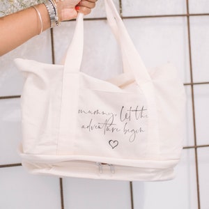 Op de afbeelding: Een witte canvas tote bag met een ritssluiting. De tas heeft een zwarte inscriptie met de tekst "mummy, let the adventure begin" met een klein hartje onder de tekst.